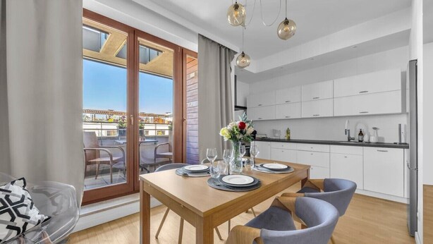 A modern kitchen with white cabinets, a dining area with a wooden table and chairs, and a balcony with outdoor seating. - Warszawa Wilanów Area Apartment — Balcony & Parking (Warszawa)