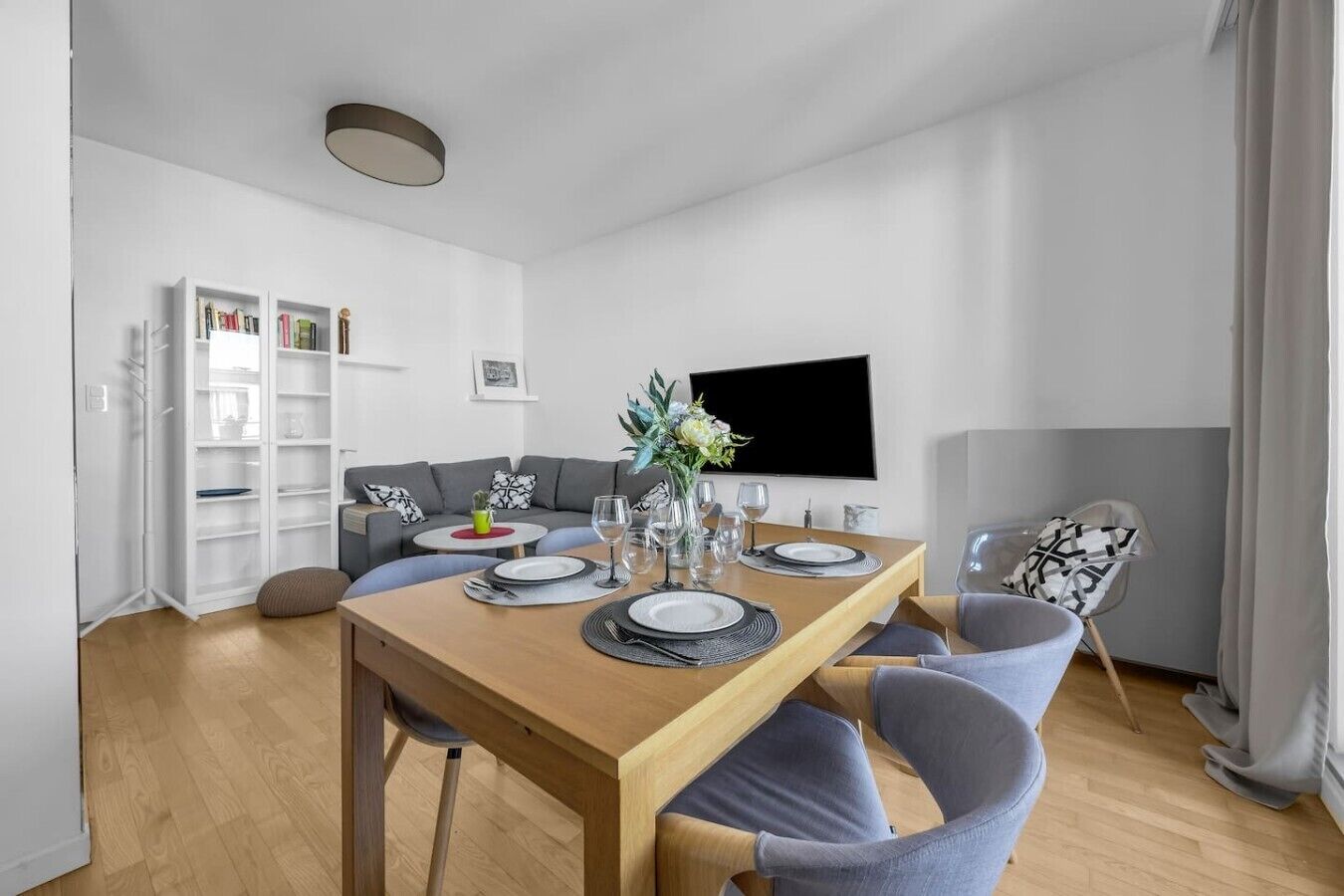 Een moderne woonkamer met een eettafel gedekt voor twee personen, een grijze bank, een aan de muur bevestigde televisie en een witte boekenkast.