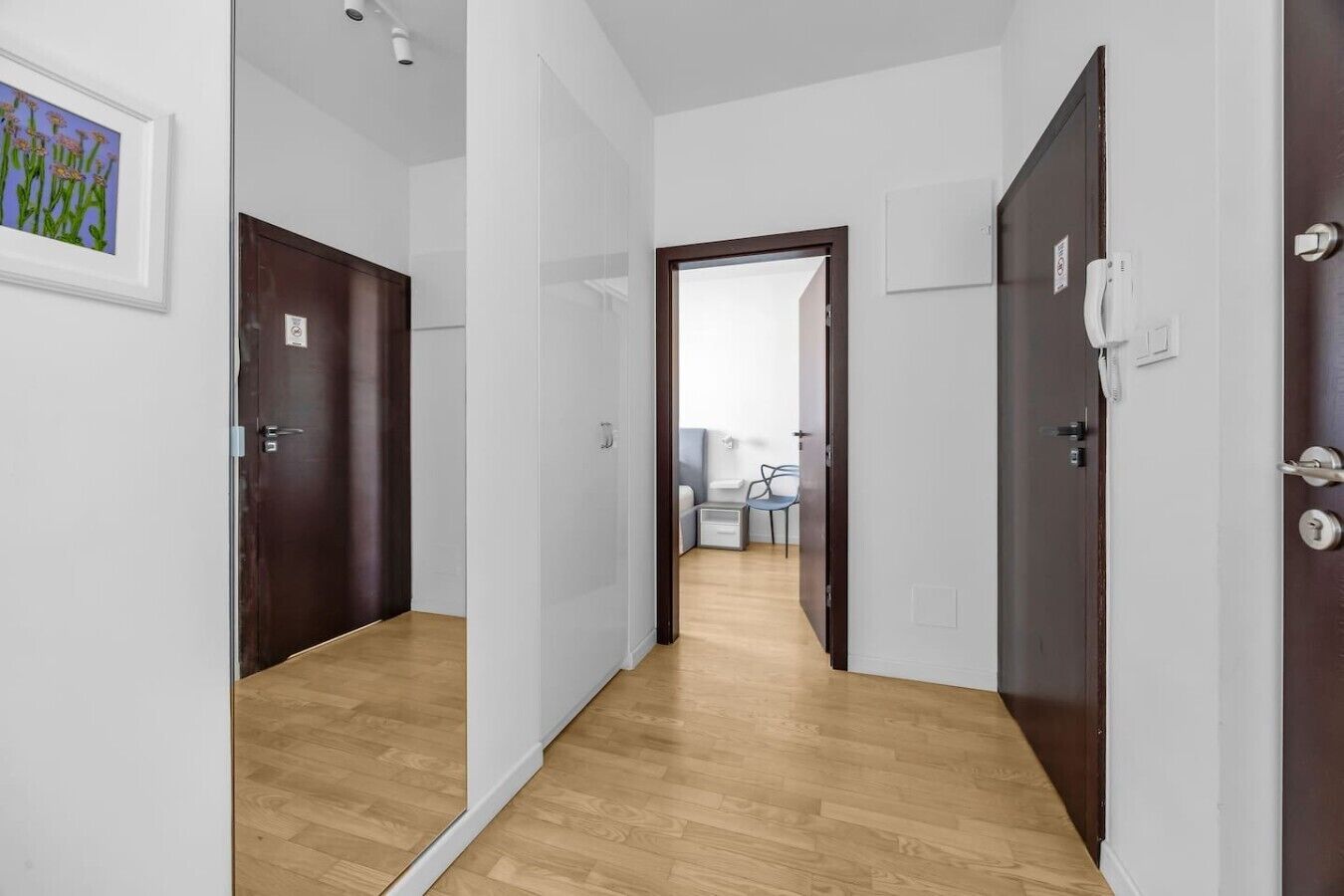 Un pasillo con piso de madera, paredes blancas y una puerta que da a una habitación con cama, silla y una mesita.