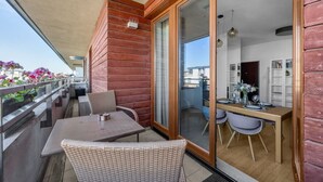A balcony with wicker furniture, a dining area with a table and chairs, and a sliding glass door leading to an interior space with a bookshelf. - Warszawa Wilanów Area Apartment — Balcony & Parking (Warszawa)