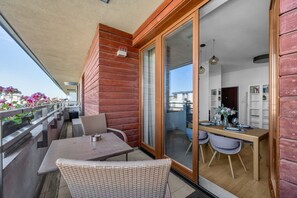 Ein Balkon mit Korbmöbeln, eine Essecke mit Tisch und Stühlen sowie eine Glastür, die zu einem Innenraum mit Bücherregal führt.