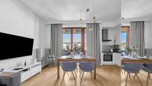 A modern living room with a flat-screen TV, a dining area with a table set for four, and a kitchen in the background. - Warszawa Wilanów Area Apartment — Balcony & Parking (Warszawa)