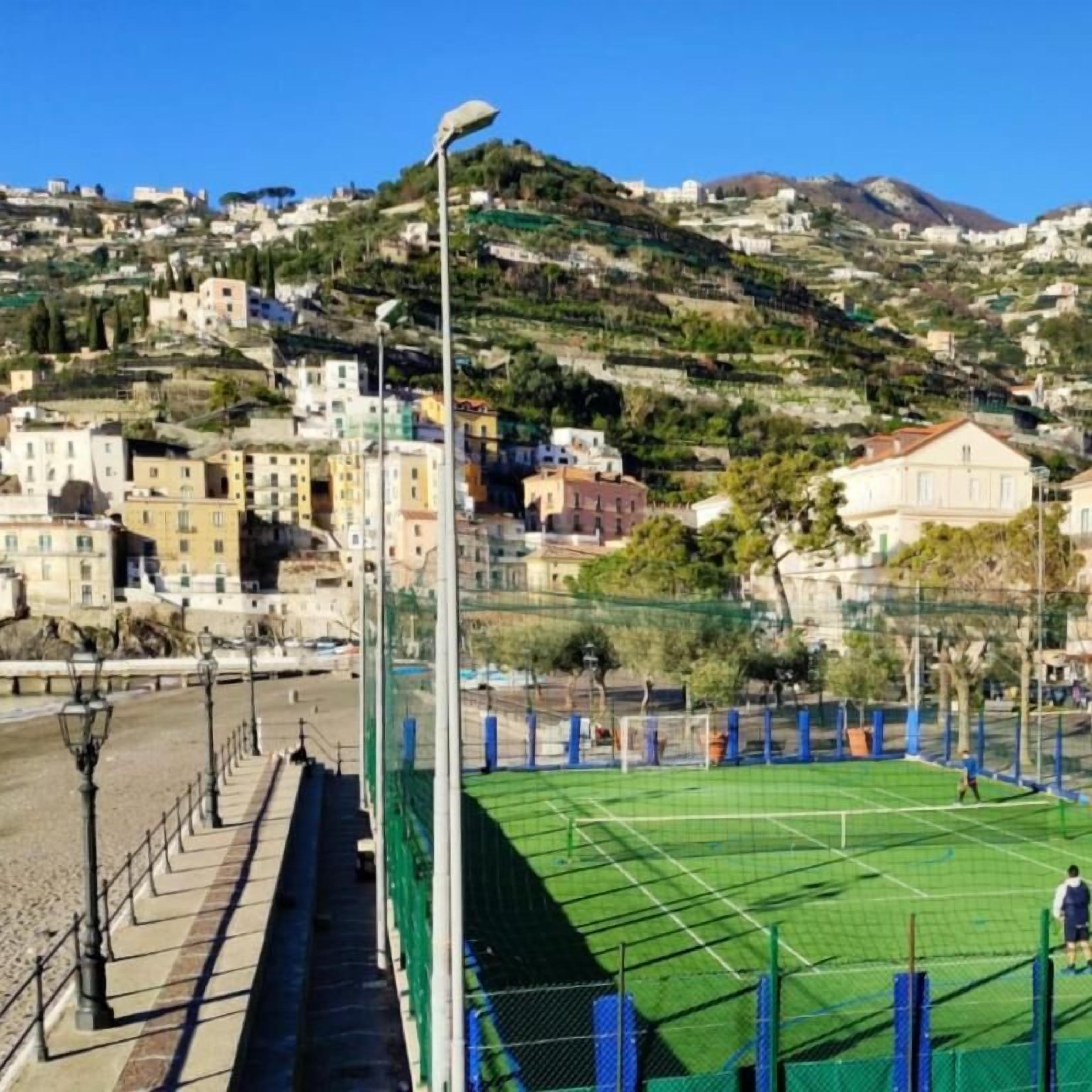 Ein Tennisplatz mit Netz, umgeben von einem grünen Zaun, mit Gebäuden im Hintergrund.