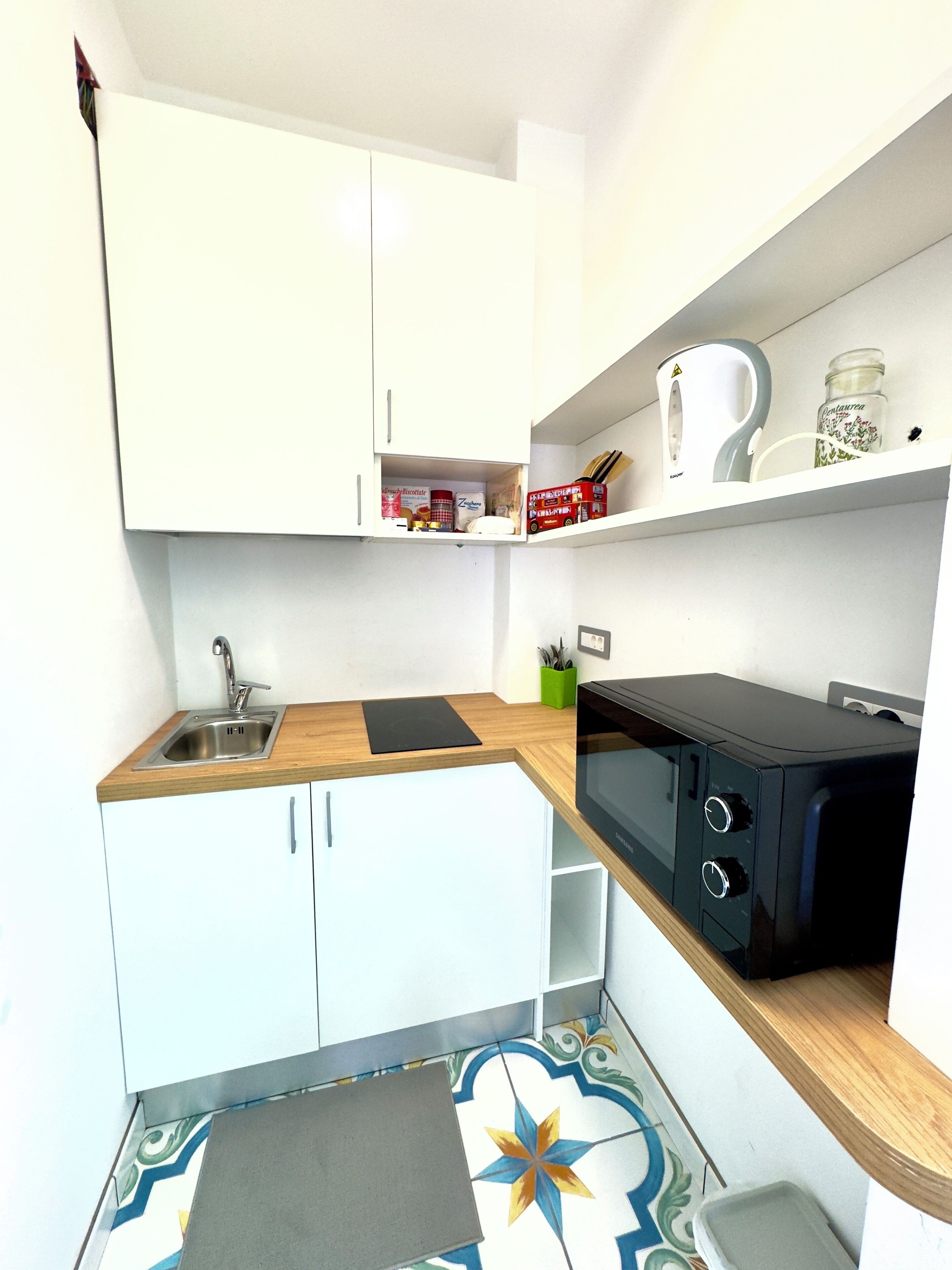 Cocina compacta con armarios blancos, un fregadero, microondas y una pequeña estantería con utensilios de cocina.