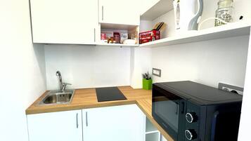 Cocina compacta con armarios blancos, un fregadero, microondas y una pequeña estantería con utensilios de cocina.