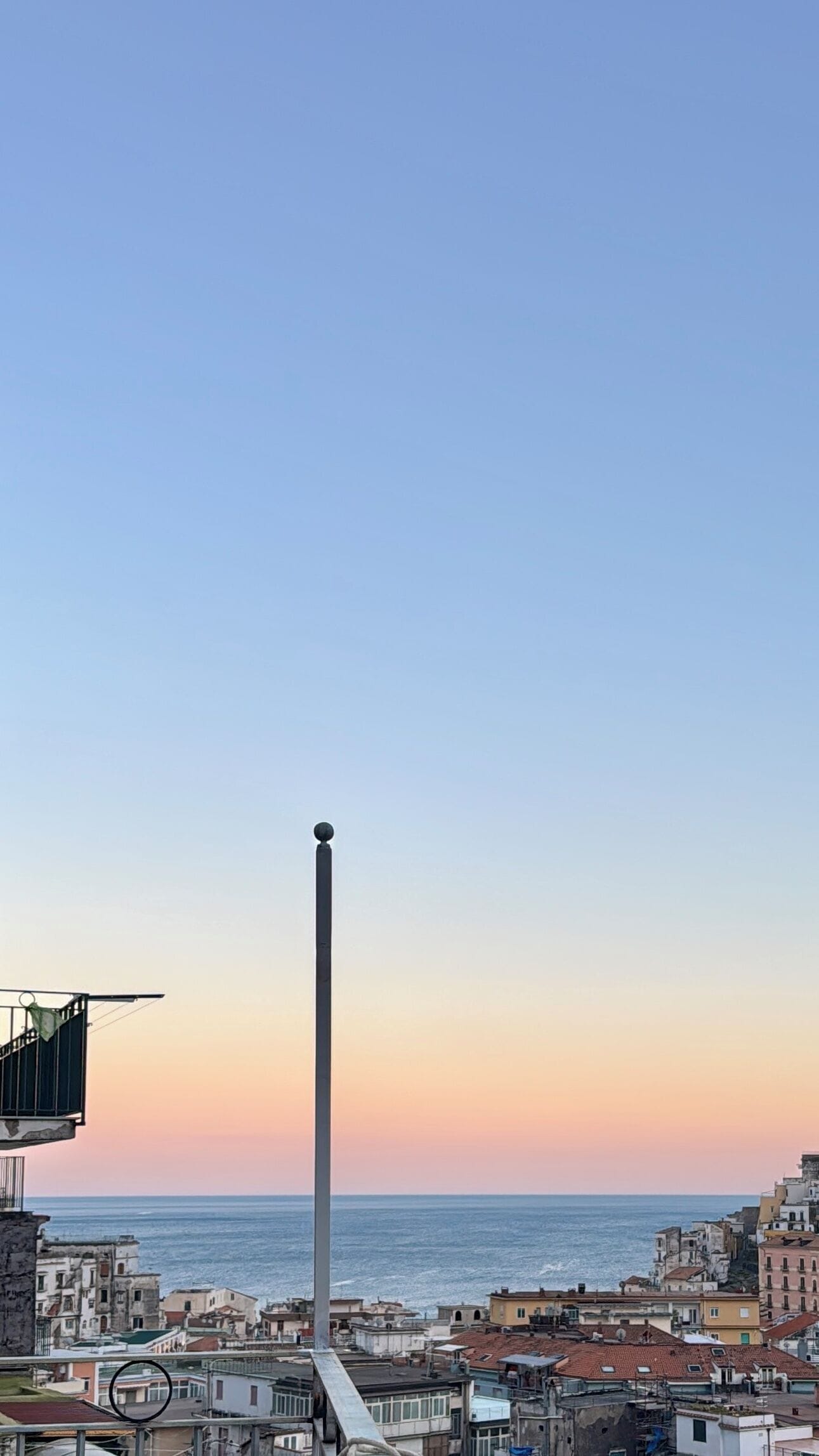 A tall, narrow structure with a spherical top, overlooking a coastal cityscape with buildings and the sea.