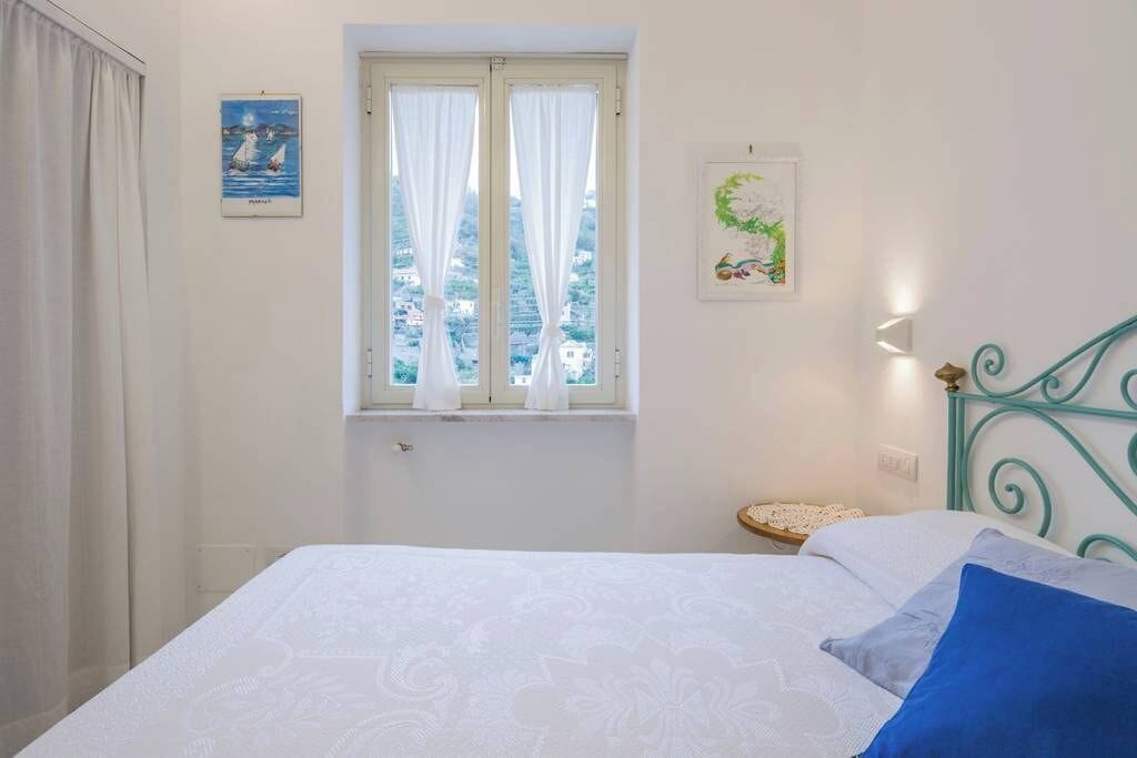 Une chambre à coucher avec un lit, une fenêtre avec des rideaux, un luminaire fixé au mur et une petite table sur laquelle se trouve un objet décoratif.