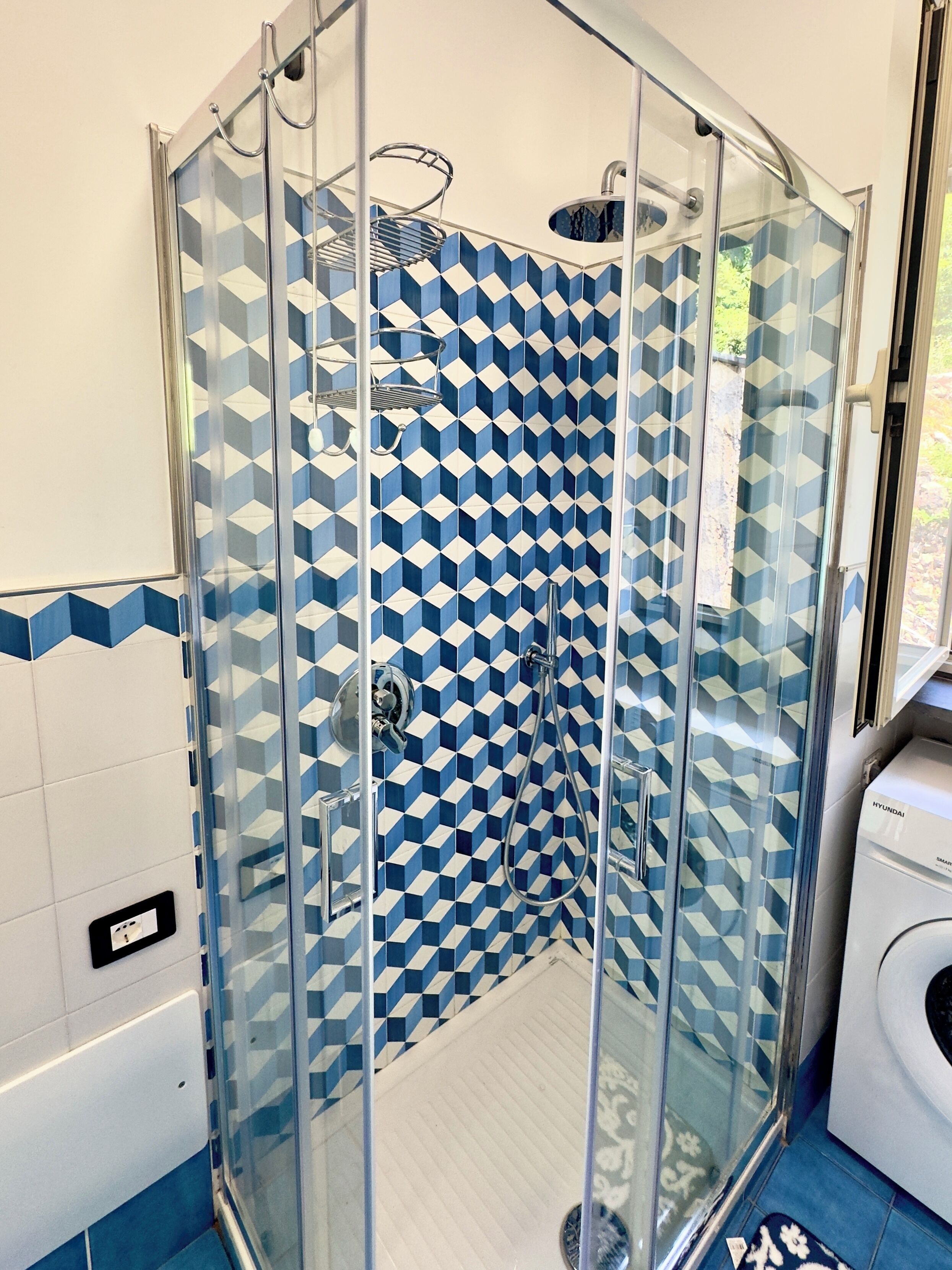A glass-enclosed shower with a geometric tile pattern on the walls, a visible water outlet, and a washing machine in the adjacent room.