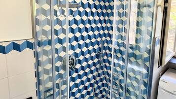 A glass-enclosed shower with a geometric tile pattern on the walls, a visible water outlet, and a washing machine in the adjacent room.