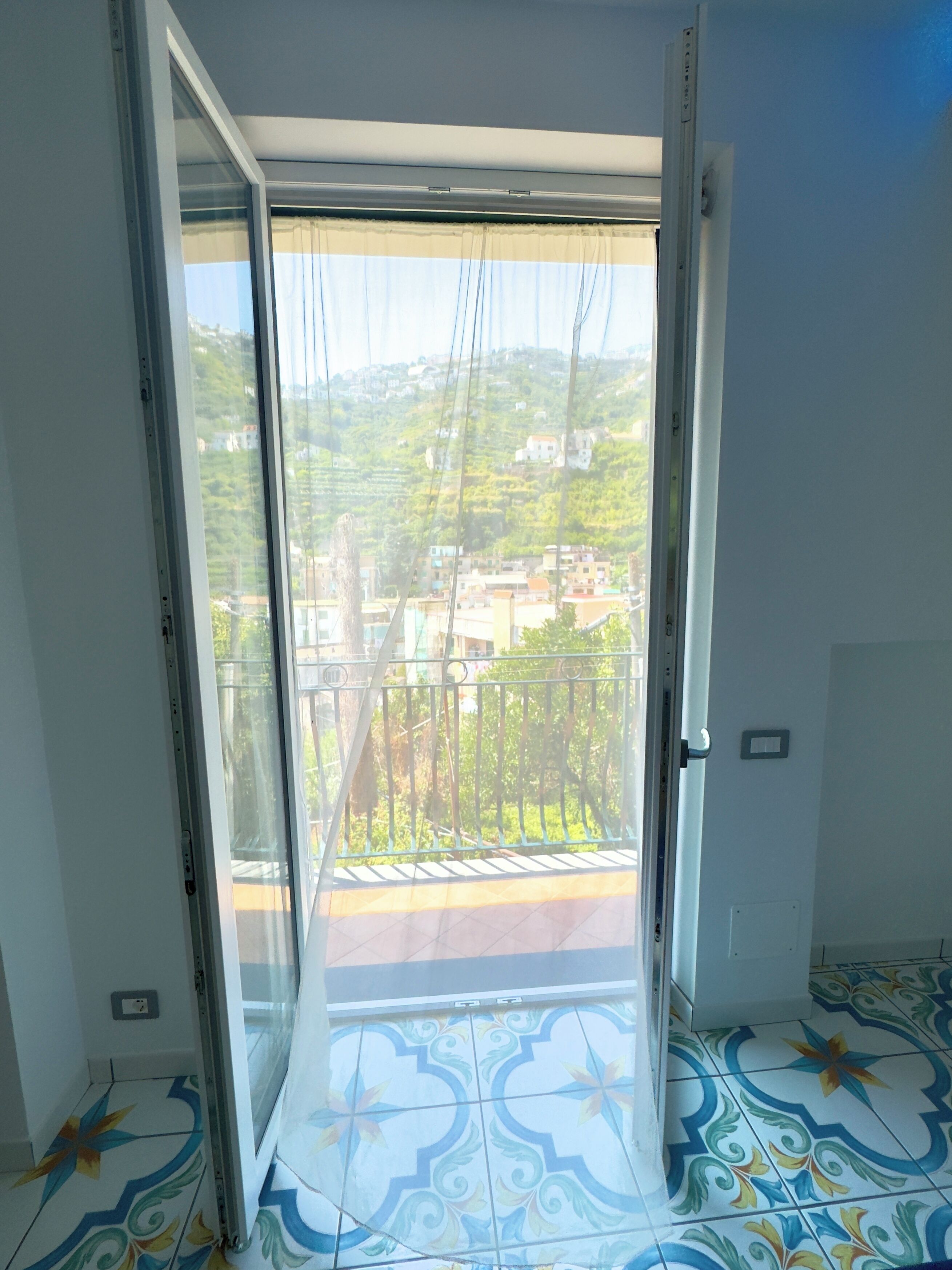 A room with a sliding glass door leading to a balcony with a view of a hillside and buildings.