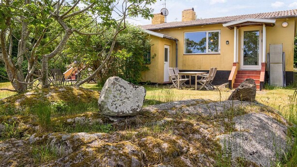A house with a yellow exterior, a large window, and a door. There is a table and chairs outside, and a rock in the foreground. - 5 Person Holiday Home in Allinge (Allinge)