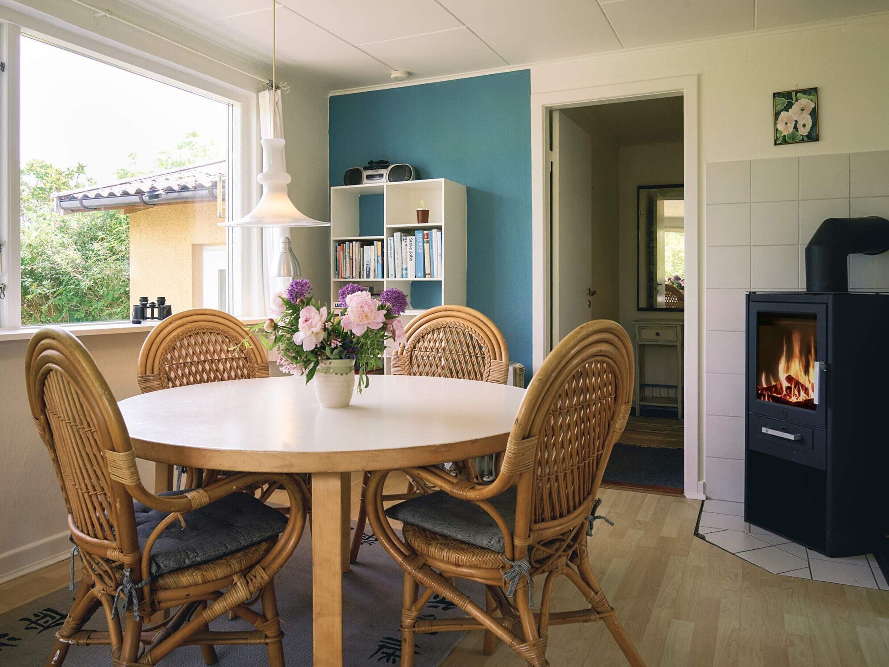 A dining area with a round table, four wicker chairs, a vase with flowers, and a bookshelf in the background.