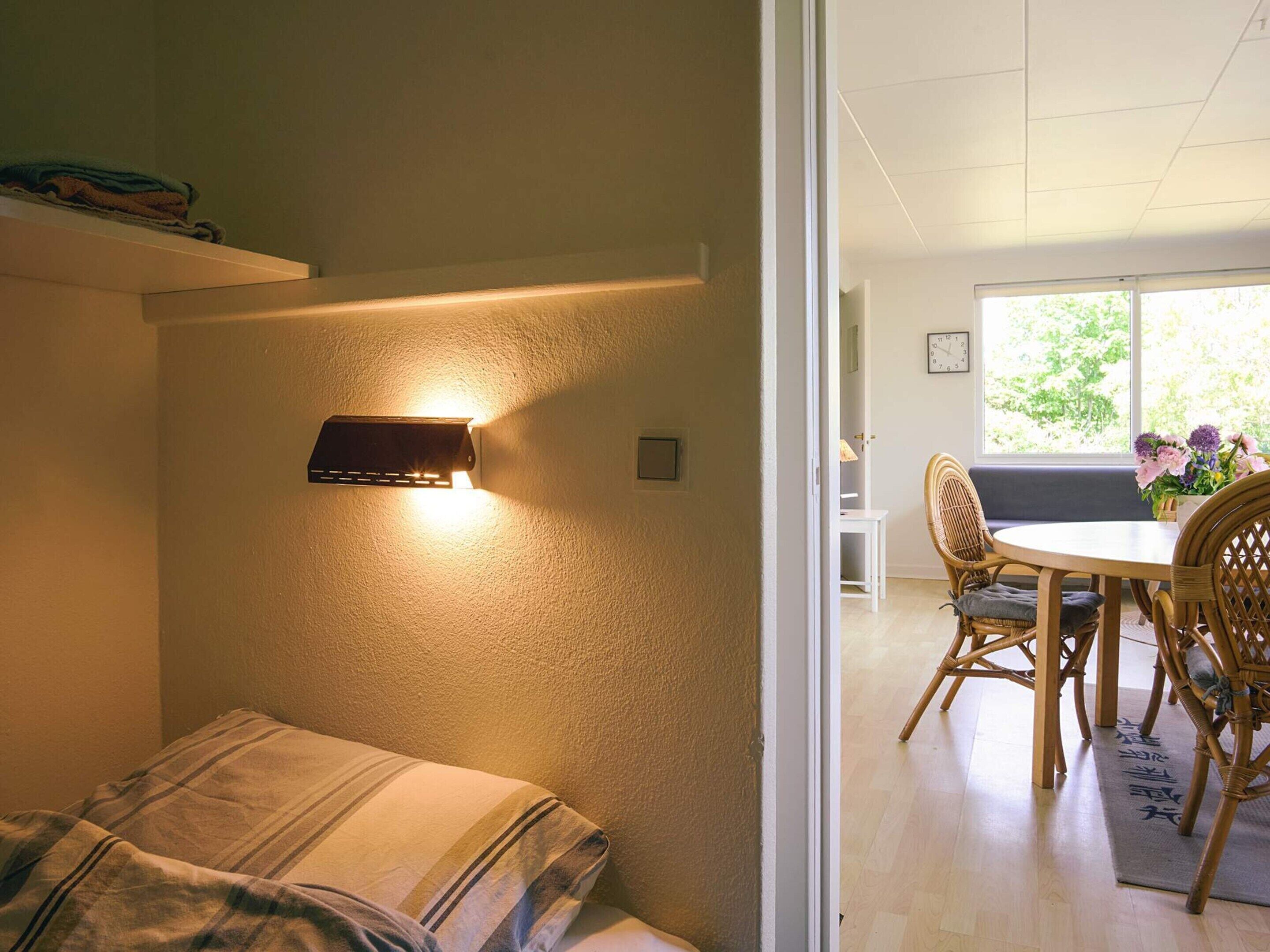 A room with a bed, a wall-mounted light fixture, a dining area with chairs and a table, and a window with natural light.