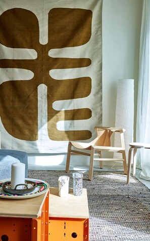 Una habitación con un tapiz abstracto grande, dos sillas de madera, una mesa pequeña con un objeto blanco y una alfombra estampada.
