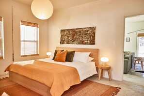 A bedroom with a large bed, two bedside tables with lamps, a cityscape artwork on the wall, and a view of a living area through an open door.