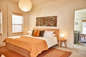 A bedroom with a large bed, two bedside tables with lamps, a cityscape artwork on the wall, and a view of a living area through an open door.