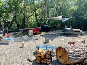 An outdoor area with a gravel ground, a pile of firewood, metal containers, and a table with chairs.