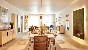 Een eethoek met een lange tafel, rieten stoelen, een hanglamp en een groot abstract schilderij aan de muur.