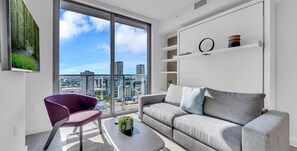 A modern living room with a grey sofa, a purple chair, and a large window offering a city view. - Yotel (Miami)