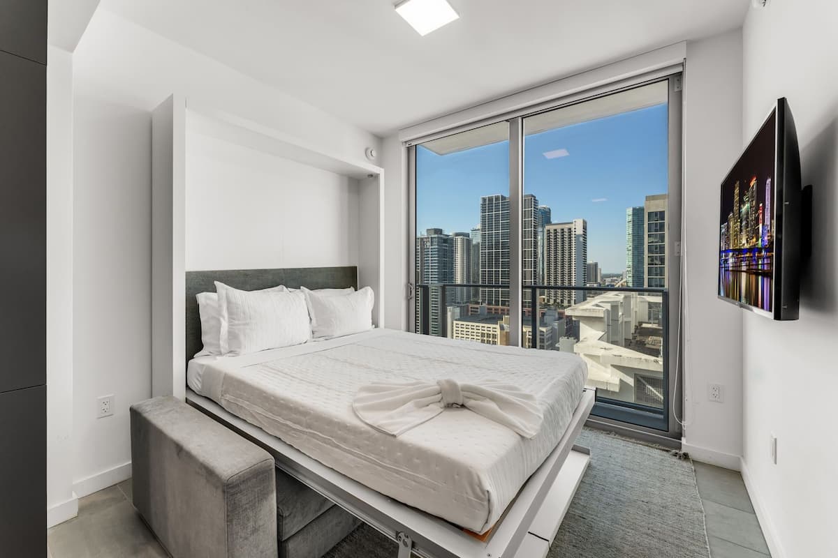 A modern bedroom with a large bed, a view of the city through a floor-to-ceiling window, and a flat-screen TV mounted on the wall.