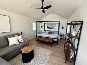 Un dormitorio con una cama con dosel, un sofá gris, una mesa de centro de madera, un taburete negro, una alfombra estampada y un estante metálico negro.