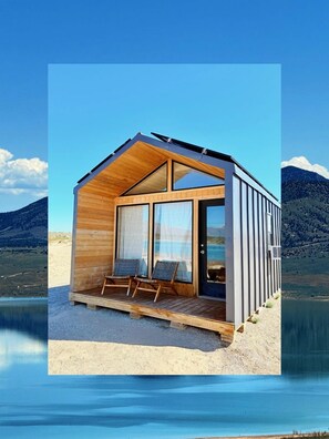 Una cabaña de madera con puerta de vidrio, dos sillas y vistas a un lago y montañas.