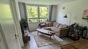 Een woonkamer met een bank, een salontafel, een houten tafel, een lamp en een raam met uitzicht op groen.