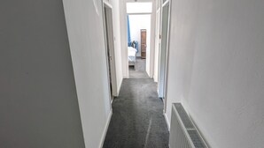 Een smalle gang met witte muren, een donker tapijt en een radiator aan de rechterkant.