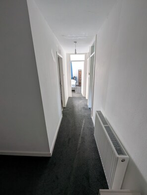 Ein schmaler Flur mit weißen Wänden, einem dunklen Teppich und einem Heizkörper auf der rechten Seite.