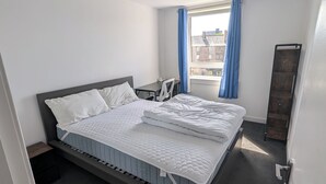Een slaapkamer met een bed, een bureau, een stoel, een raam met gordijnen en uitzicht op gebouwen.
