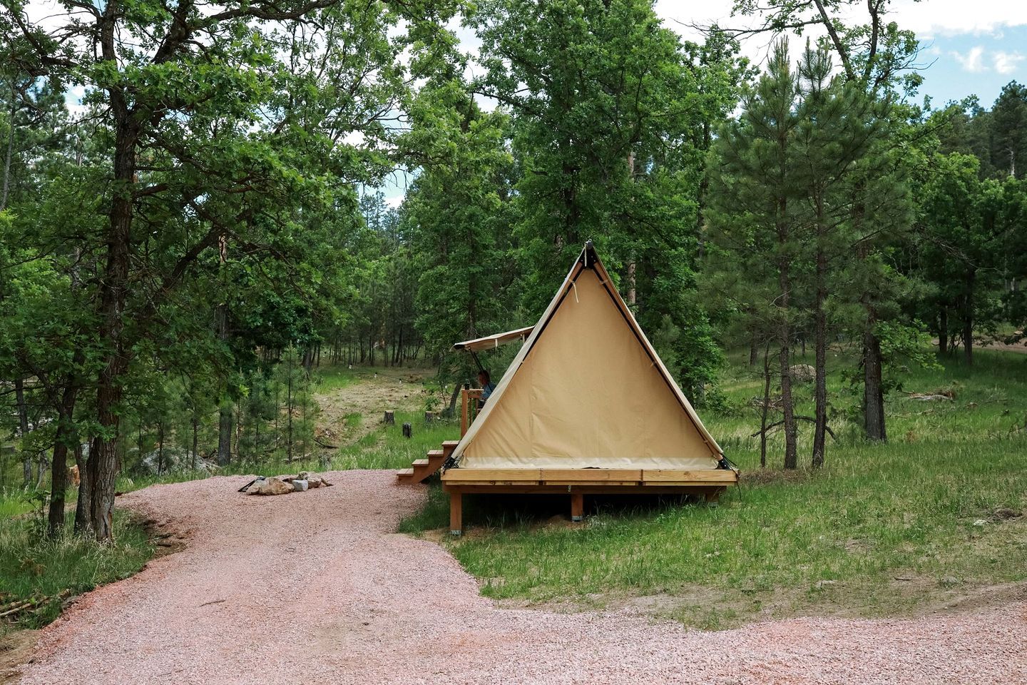 Tienda triangular con una plataforma de madera y escalones que conducen a ella, rodeada de un área boscosa.