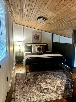 Une chambre avec un plafond en bois, un lit recouvert d’une couvre-lit à motifs, un tapis, une table de chevet avec une lampe et une œuvre d’art accrochée au mur.
