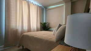 Ein Schlafzimmer mit einem Bett, einem Nachttisch mit Lampe, einer Topfpflanze und einem Fenster mit Vorhängen.