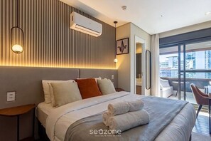 Ein Hotelzimmer mit Bett, Nachttischen, einer Klimaanlage, einer Wandleuchte und Blick auf die Stadt durch große Fenster.