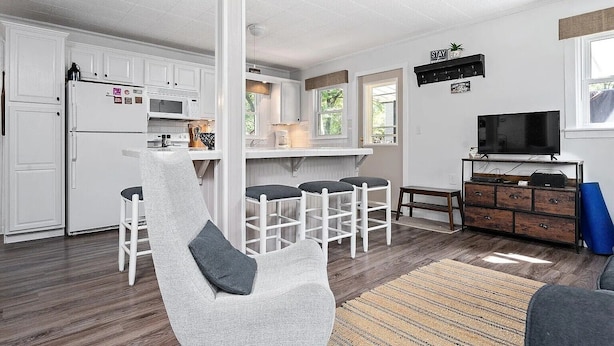 A modern kitchen with white cabinets, a refrigerator, and a microwave. There are bar stools and a television in the living area. - Round Lake | Fire Pit, Grill, Patio & Lake Access (Mecosta)