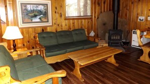 Um quarto com piso de madeira, um sofá, duas cadeiras, uma mesa, um abajur, uma lareira e uma janela.