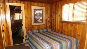 Quarto com paredes de madeira, uma cama com colcha listrada, um quadro pendurado na parede e uma janela com persianas.