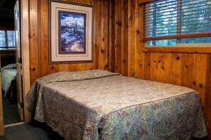 Ein Zimmer mit Holzboden, einem Bett, einem gerahmten Bild an der Wand und einem Fenster mit Jalousien.