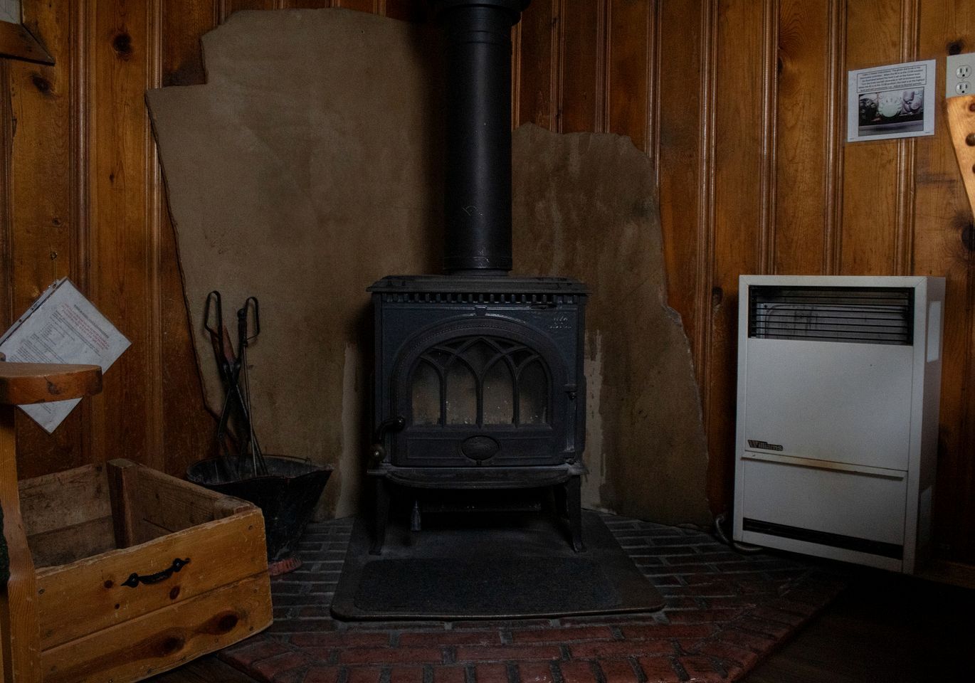 Ein Zimmer mit Kaminofen, Heizlüfter, Holzfach und Boden aus Ziegelsteinen.