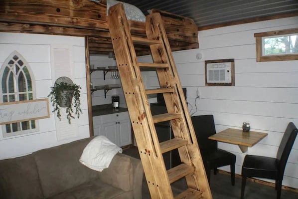 Habitació d'estil loft amb escala de fusta, petita cuina, zona de menjador i finestra amb vista.