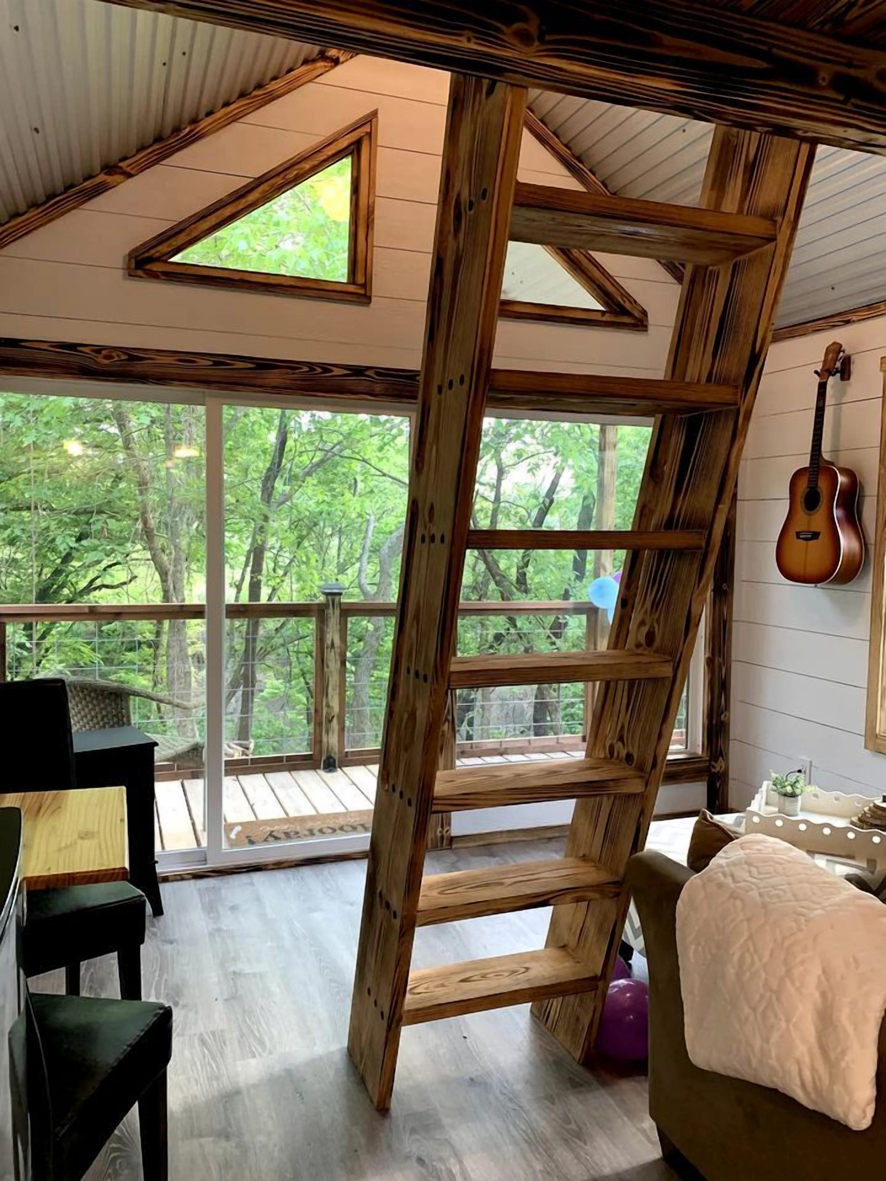A rustic wooden interior with a loft bed, a guitar, and a large window overlooking a deck.