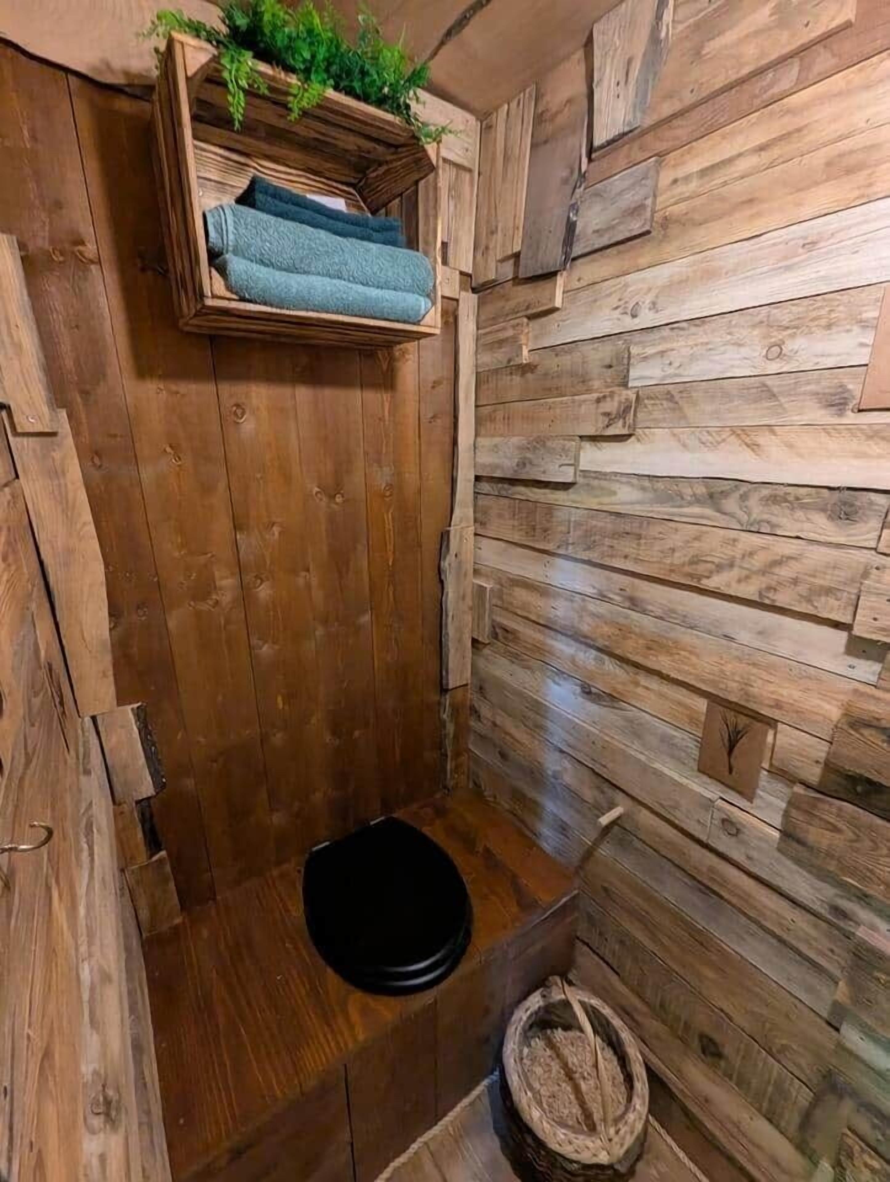 Une salle de bain rustique en bois, avec des toilettes noires, une étagère en bois où sont posées des serviettes et un panier en osier.