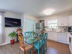 Une cuisine compacte avec des meubles blancs, une table à manger en verre et un téléviseur fixé au mur.