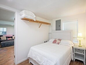 Une chambre à coucher avec un lit, une table de chevet, une étagère avec un coussin et une fenêtre avec des stores.