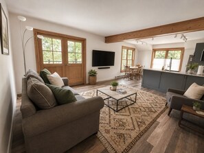 A modern living room with a sofa, coffee table, and TV. There are wooden floors, large windows, and a kitchen area.