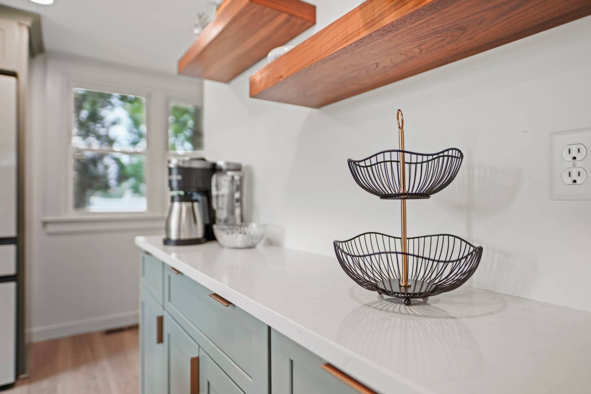 A kitchen with a white countertop, a two-tiered wire fruit basket, and a coffee maker.