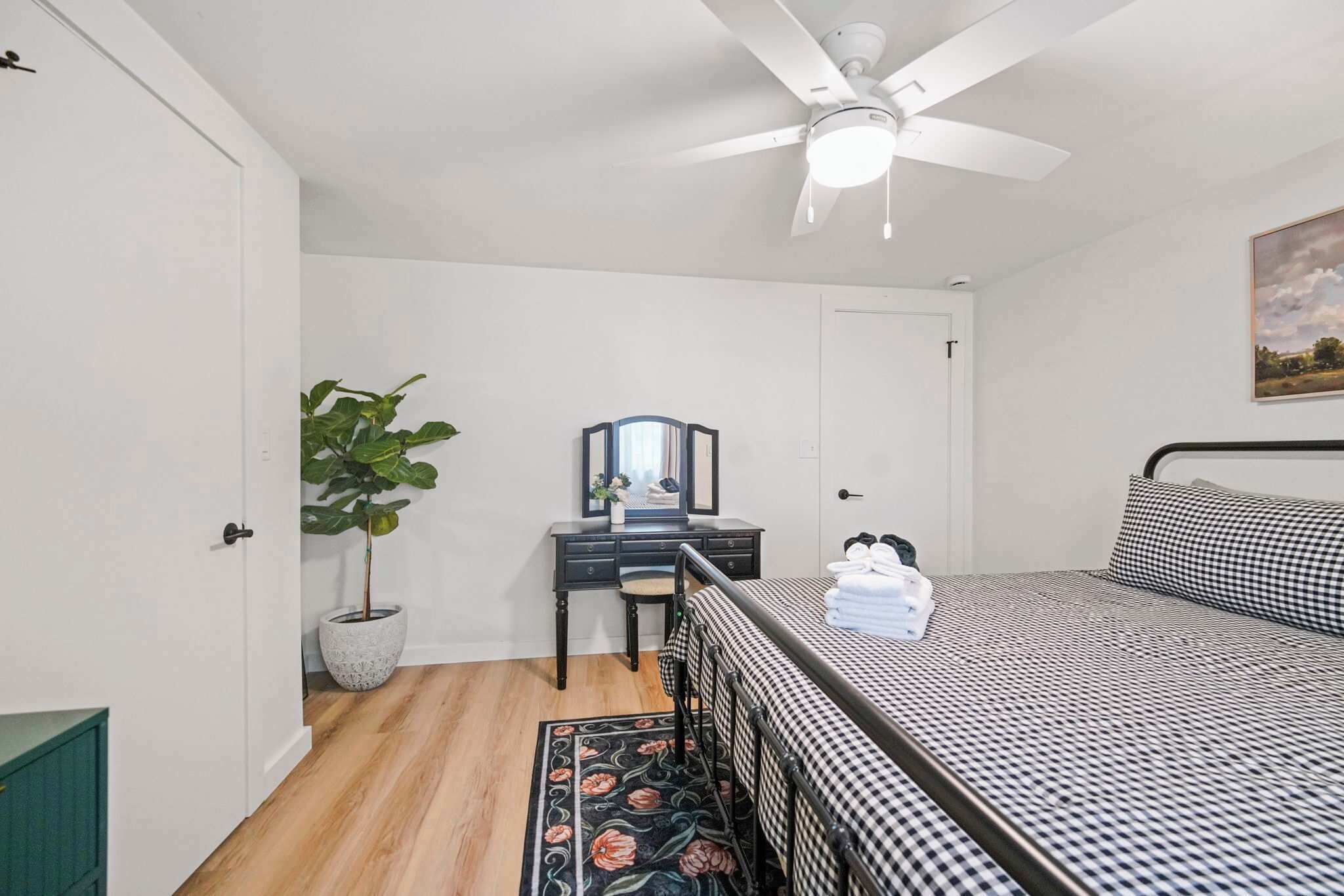 Un dormitorio con una cama, un escritorio con silla, una planta en maceta, un ventilador de techo y una puerta.