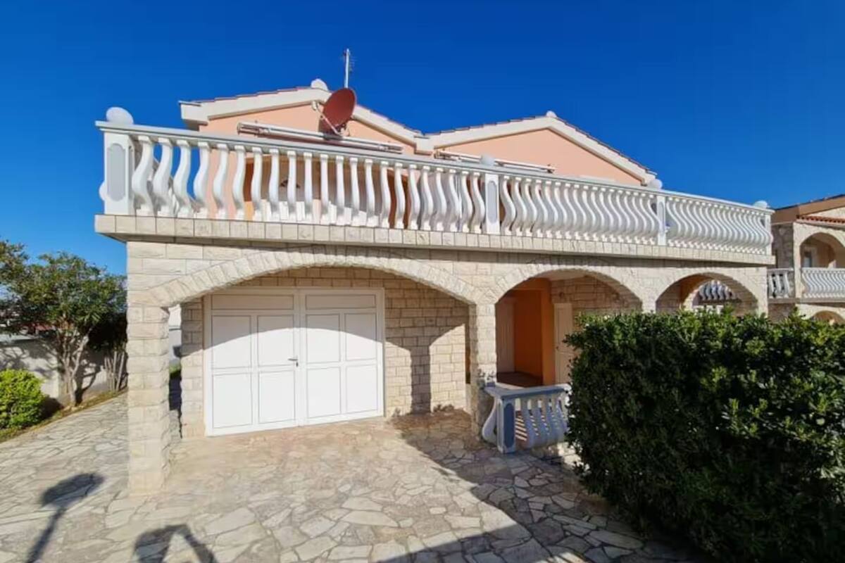 Un immeuble de deux étages avec une façade en pierre, une porte de garage blanche et un balcon doté d’une balustrade.
