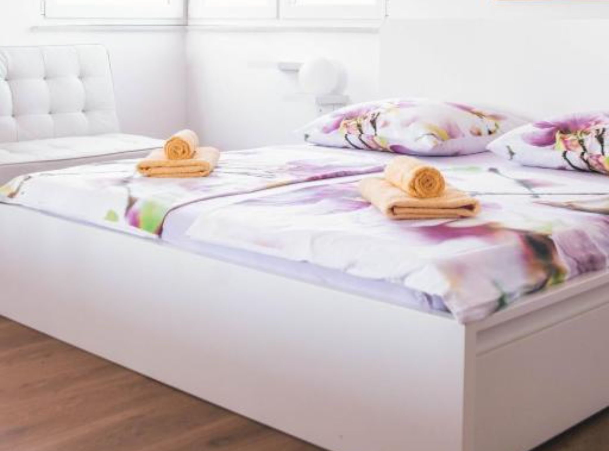 A neatly made bed with floral bedding, a white headboard, and rolled towels on the side.