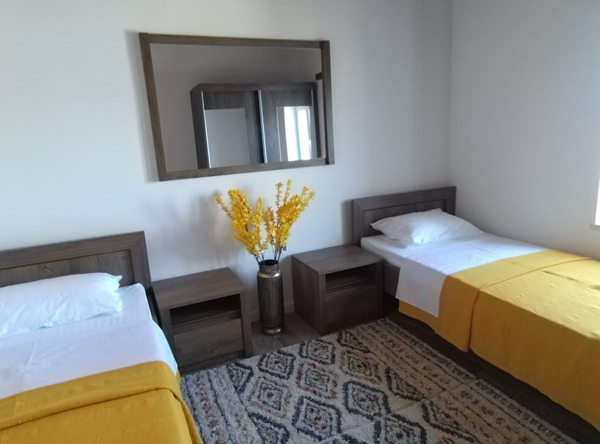 Quarto de hotel com duas camas, uma mesa de cabeceira com um vaso de flores, um tapete estampado e um espelho sobre as camas.